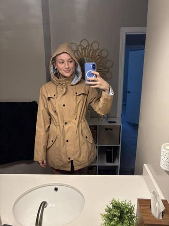 Person in a hooded tan jacket takes a mirror selfie in a bathroom, showcasing the jacket's style and fit