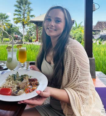 Person seated at an outdoor restaurant, smiling, holding a plate with assorted food, including vegetables and meats, with a lush greenery background