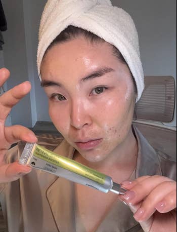Person with towel on head applies tube skincare product on dewy face, wearing robe