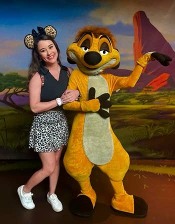 Person posing with a Timon mascot from The Lion King. The person is wearing Minnie Mouse ears, a leopard print skirt, and white sneakers