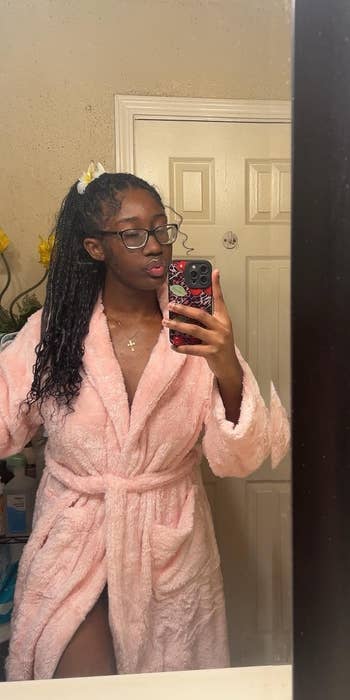 Person in a bathroom mirror selfie wearing a fluffy bathrobe and glasses, posing with hair tied back
