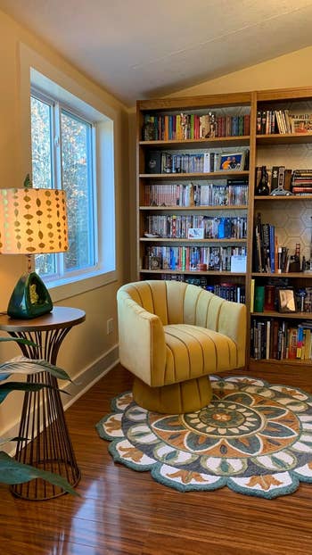 A cozy reading nook features a plush, round chair in front of a bookcase filled with various books, DVDs, and decor items. A side table with a lamp is nearby