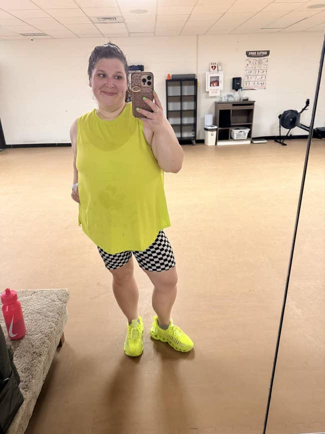 Person taking a mirror selfie in a fitness studio, wearing a sleeveless top, checkered shorts, and sneakers, standing next to fitness gear