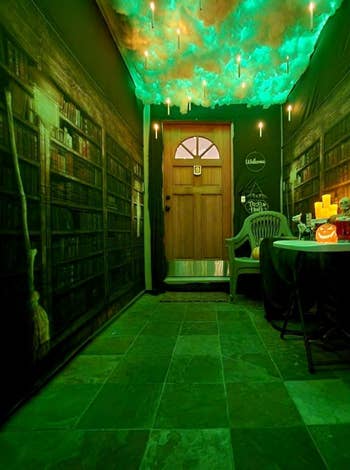 Hallway decorated with floating candles, cloud-like ceiling, and wallpaper of bookshelves. A table with Halloween-themed items and a pumpkin sits near the door