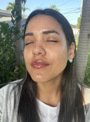 Person with long hair enjoys the sun, eyes closed, wearing a casual top and earrings, exemplifying a serene moment outdoors