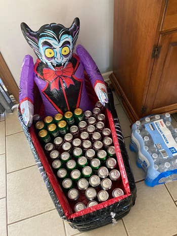 Inflatable vampire decoration holding assorted canned drinks in a bathtub-shaped container next to a pack of bottled water
