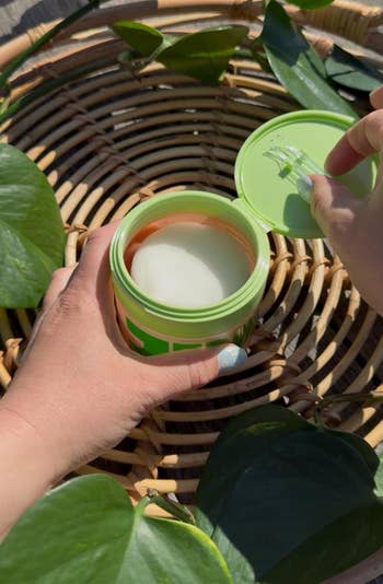 reviewer opening a skincare jar with a toner pads inside, surrounded by greenery on a woven tray