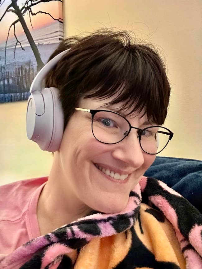 Person smiling, wearing headphones and glasses, wrapped in a patterned blanket, in a cozy setting