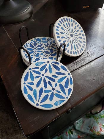 Set of four decorative coasters with blue floral and geometric patterns, placed in a black metal holder on a dark wooden surface