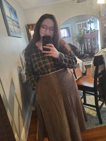 A person in a plaid shirt and long skirt takes a mirror selfie in a cozy home setting with dining area visible in the background