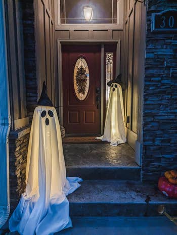 Two ghost decorations made of white fabric and hats, with glowing lights inside, placed on a porch beside a door with a fall wreath