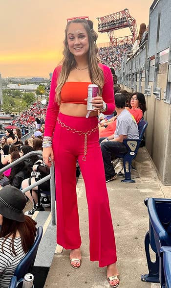 Person at a stadium, wearing a stylish orange and red outfit with gold chain belt, holding a drink. Stadium seats and attendees in the background