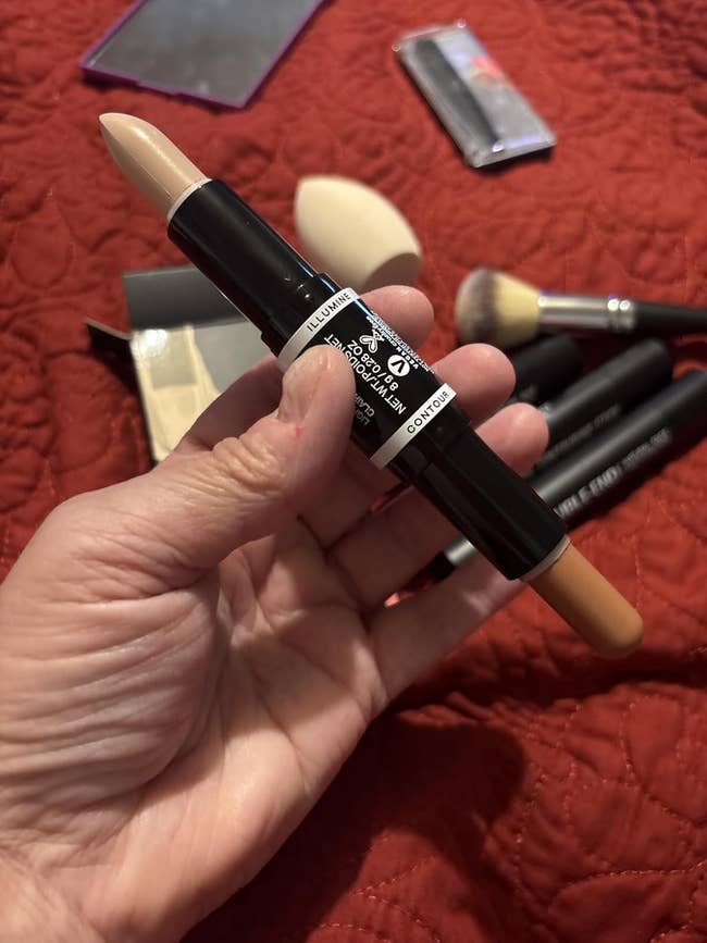 Double-ended foundation stick held in hand; makeup brushes and sponge are in the background