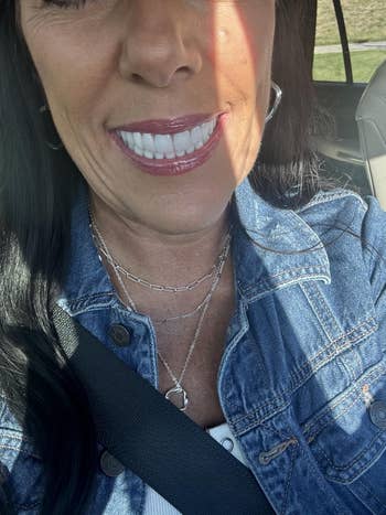 Woman wearing a denim jacket and layered silver necklaces, smiling in a car, sunlight on her face