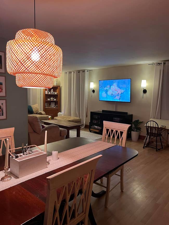 Modern dining room with wooden table, chairs, decorative ceiling light, and cozy living area with a TV and furniture in the background
