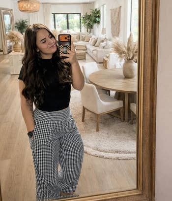 Person taking a mirror selfie in a stylish, modern living room with neutral decor and checkerboard pattern pants