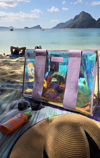 Beach scene with a transparent Case-Mate tote, sunscreen, sunglasses, and a straw hat on a colorful towel. Scenic view of sea and mountains in background