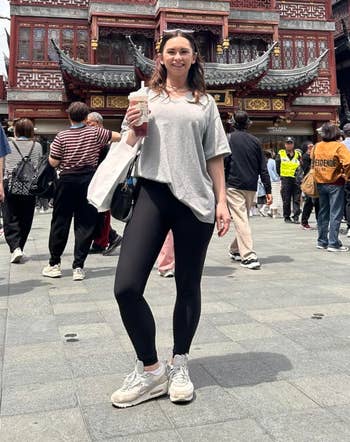 Person in casual clothes holding a drink, standing in front of a traditional Asian architectural building with a busy crowd surrounding