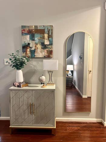 Living room setup with a geometric cabinet, a potted plant, a globe, a lamp, an abstract wall painting, and an arched mirror reflecting a lamp and staircase
