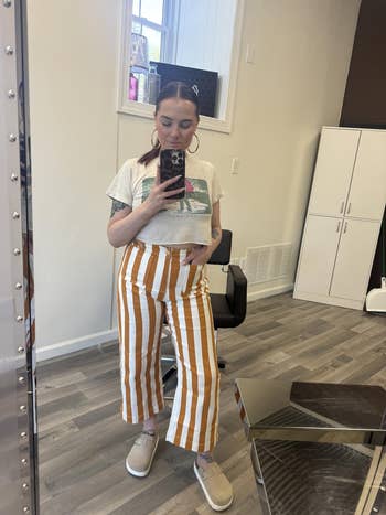 Person in a mirror selfie wearing a cropped graphic tee and striped wide-leg pants. Standing in a casual setting