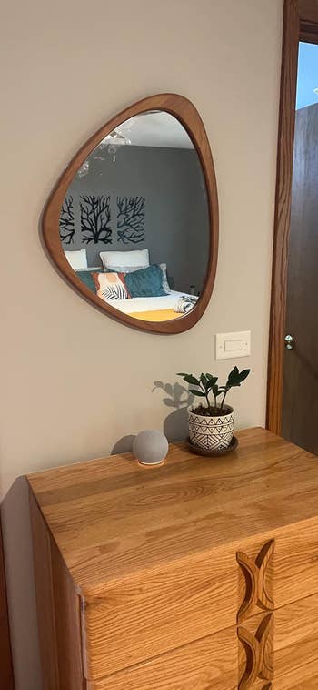 Wood-framed triangular mirror above a wooden dresser with a potted plant and a round decorative item, reflecting a bedroom with a tree-patterned headboard