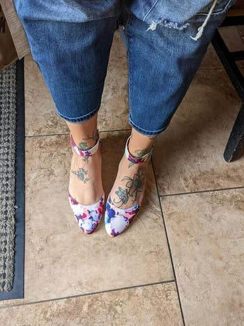 Person wearing ankle-strap floral heels, paired with rolled-up jeans, showcasing ankle tattoos on a tiled floor