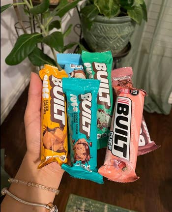 A hand holds various Built protein bars, including flavors like Salted Caramel, Cookie Dough, and Strawberry, displayed in a casual home setting