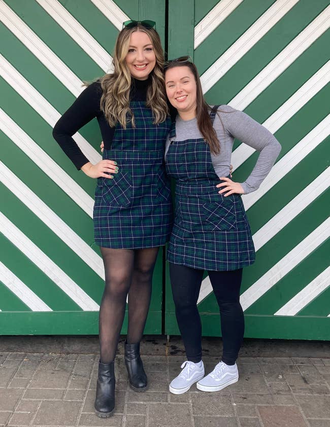 Two people smiling, wearing plaid dresses over tops, one in boots and the other in sneakers, standing in front of a green striped background