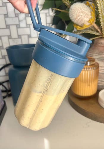 A hand holds a blue-lidded blender bottle filled with a smoothie. Kitchen items are visible in the background