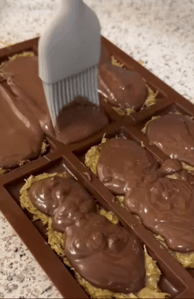 A brush coats melted chocolate over homemade granola bars in a silicone mold on a kitchen counter