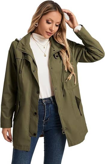 Model in casual outfit modeling an olive-green jacket, white top, and blue jeans. She wears layered necklaces, suitable for a shopping feature