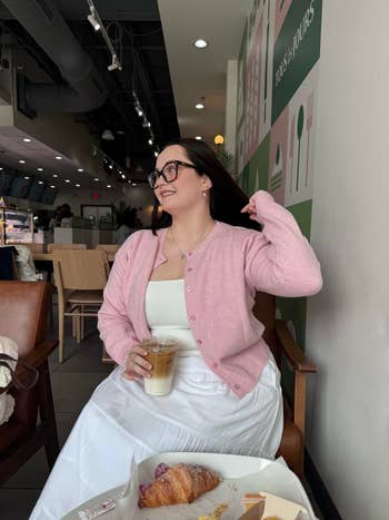 Person in coffee shop wearing a cardigan and glasses, holding a beverage. Croissant and pastry box on table. Casual, relaxed setting