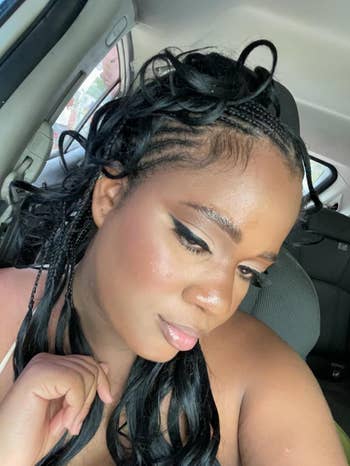 Person in a car with styled braids and winged eyeliner, gently touching their collarbone with a thoughtful expression