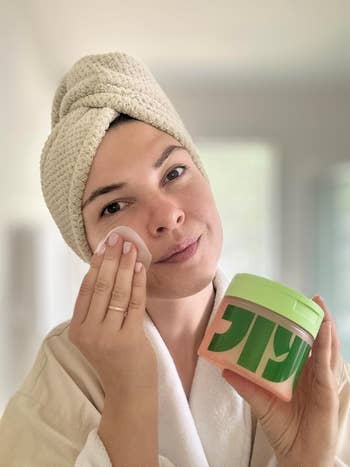reviewer with a towel wrapped around their head applies skincare product using a cotton pad while holding a jar