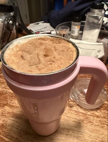 Large pink insulated tumbler filled with a cinnamon-topped drink, placed on a wooden table surrounded by kitchen items