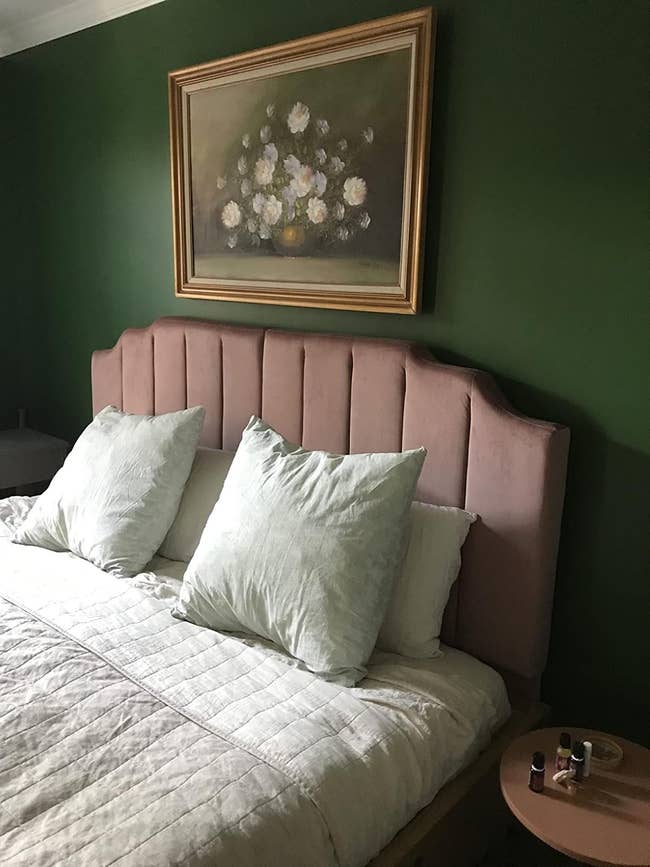 Bedroom with a padded headboard, pillows, a quilted bedspread, a small round bedside table with nail polish, and a framed floral painting on the wall