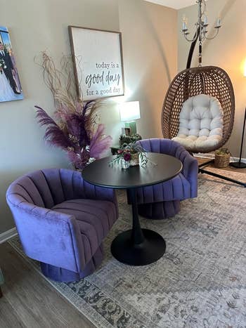 Two purple velvet chairs, a black round table with a floral centerpiece, a wicker egg chair, and a wall sign saying,