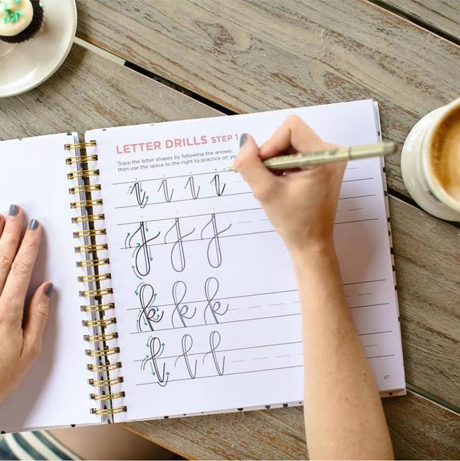 Hand practicing cursive lettering in a spiral-bound workbook beside a coffee cup and cupcake