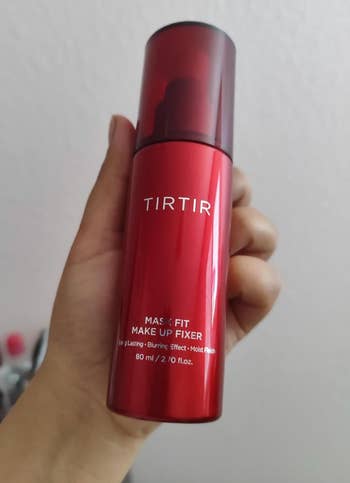 Hand holding a red bottle of TIRTIR Mask Fit Makeup Fixer with text highlighting long-lasting, blurring effect, and moisture benefits
