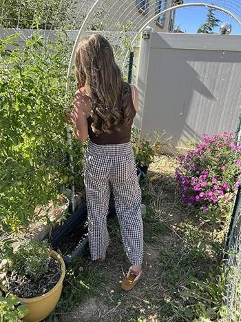 Person with long hair tends to plants in a garden, wearing a sleeveless top and checkered pants, with a pot and flowers nearby