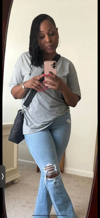 Person in casual outfit with a gray top and ripped jeans holding a phone and posing in front of a mirror, wearing a crossbody bag