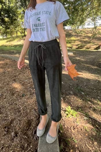 Person wearing a casual T-shirt and pants stands on a wooden edge outdoors holding a leaf