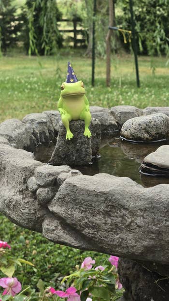 A toy frog wearing a wizard hat sits on a rock in a garden water feature
