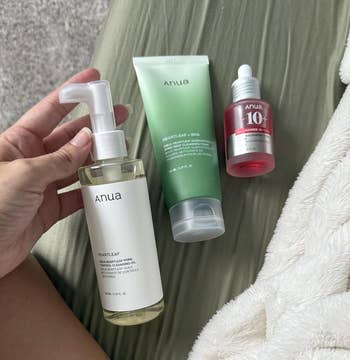 Three Anua skincare products: a pump bottle, a green tube, and a red serum bottle, displayed on a bed with a plush blanket