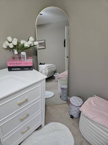 A bedroom corner features a dresser with books and a vase of white tulips, a large arched mirror, an ottoman, and a glimpse of a bed reflected in the mirror