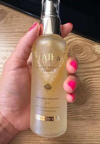 A hand holding a bottle of d'Alba First Spray Serum, a skincare product promoting hydrated and radiant skin