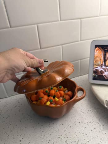 Hand lifting lid of pumpkin-shaped dish filled with candy pumpkins on a kitchen counter next to a tablet