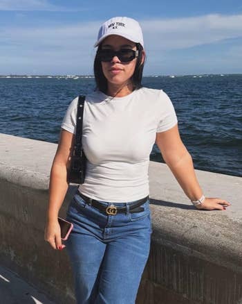 Person wearing a white cap and sunglasses, in a casual white top and jeans, stands by a waterfront