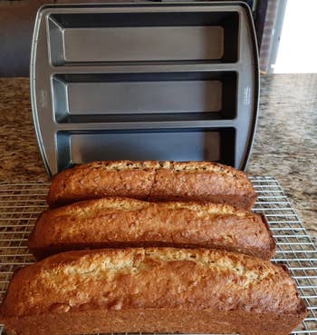 Three freshly baked loaves of banana bread cooling on a rack, with a nonstick loaf pan in the background