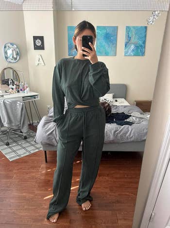 Person in a bedroom mirror selfie wearing a cozy loungewear set, with a twisted-front top and relaxed pants. Room has casual decor